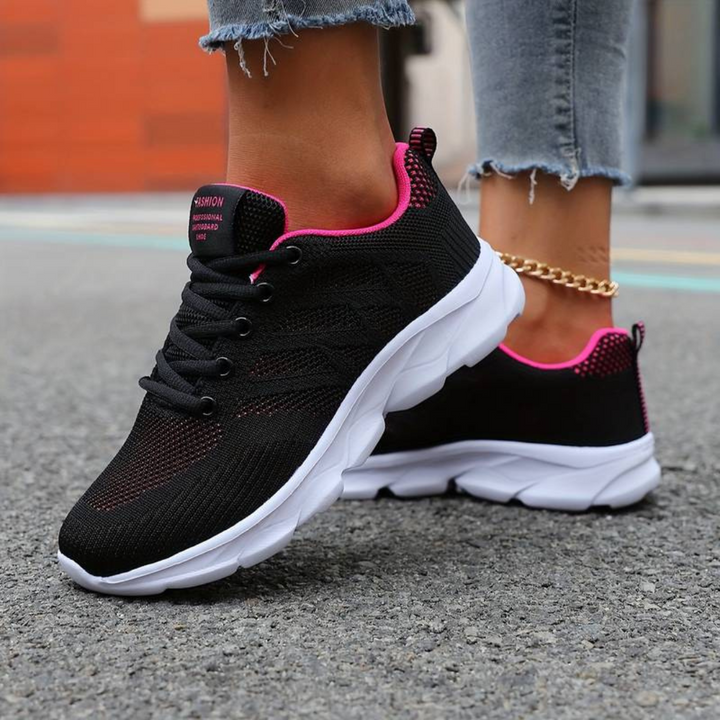 Schwarze Damen-Sneaker mit pinken Akzenten und weißer Sohle auf Asphalt, getragen von Person mit Jeans und goldener Knöchelkette.