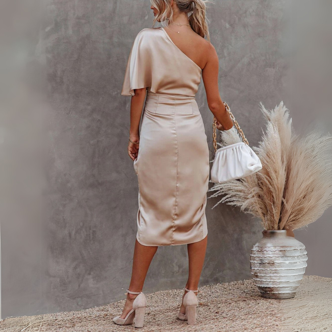 Frau in eleganten, einärmeligen, beigen Satin-Kleid mit weißer Handtasche, steht vor grauer Wand und Pampasgras in Vase. Fashion, Abendmode, Damenkleidung.