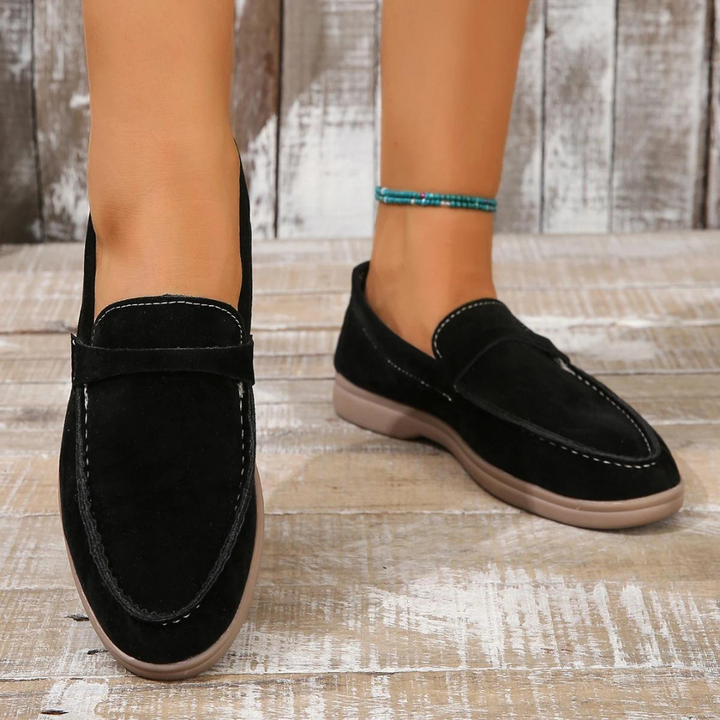 Schwarze Wildleder-Loafer auf Holzboden, getragen von einer Person mit türkisfarbenem Fußkettchen. Modische Damen-Schuhe, bequem und elegant.