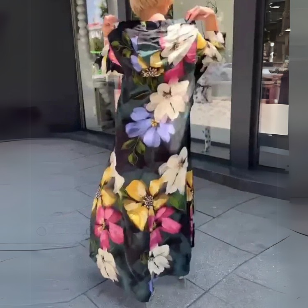 Frau in langem, geblümtem Kleid auf Gehweg vor Schaufenster. Buntes Blumenmuster, modisch, sommerlich. Ideal für urbane Mode, Streetstyle.