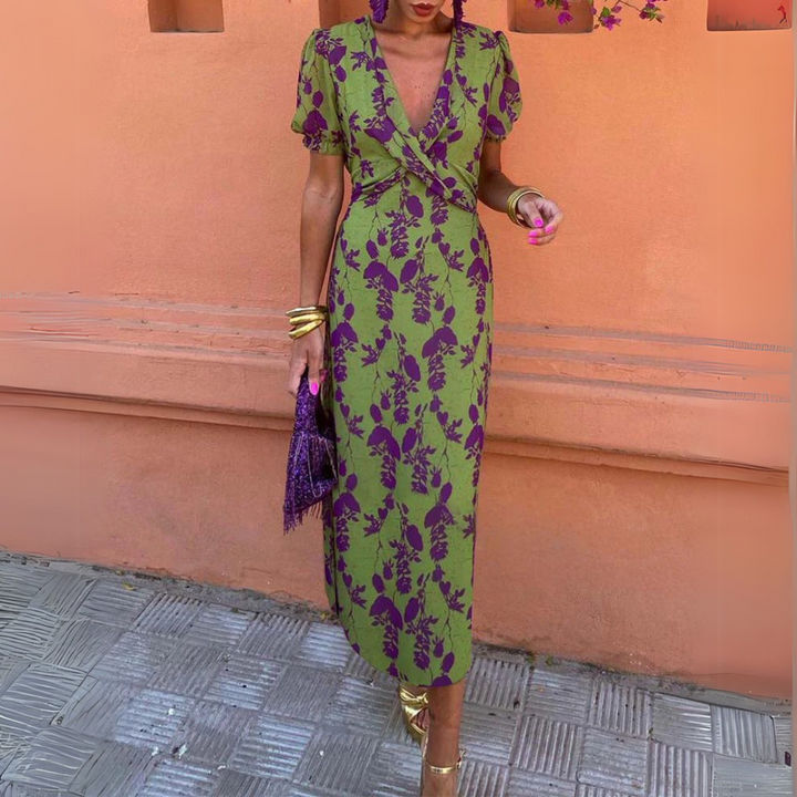 Frau in grünem, geblümtem Kleid vor orangefarbener Wand, hält lila Tasche. Sommermode, elegantes Design, florales Muster, Streetstyle.