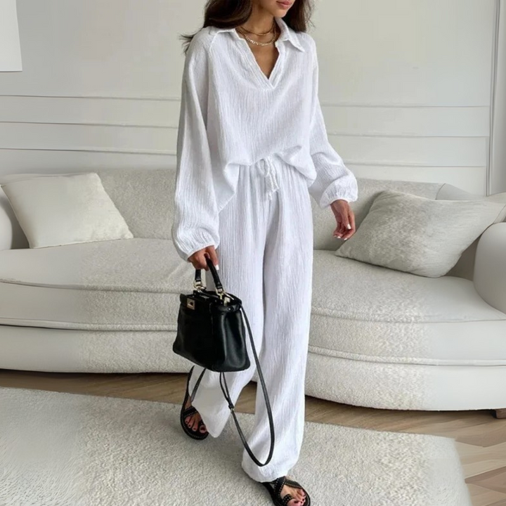 Frau in weißem Leinenanzug mit schwarzer Handtasche, steht vor cremefarbenem Sofa. Elegante, minimalistische Mode, lässiger Stil.
