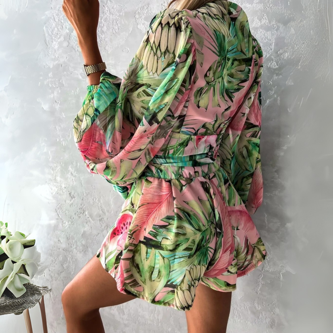 Frau in tropischem, geblümtem Kleid mit Ananas- und Blattmuster, posiert vor grauer Wand. Sommermode, farbenfrohes Design, stilvoller Look.