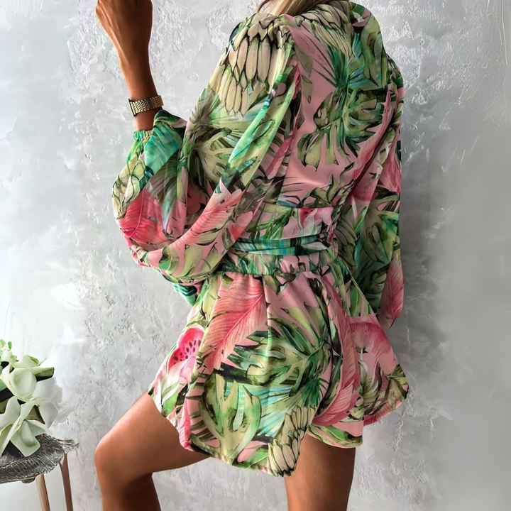 Frau in tropischem, geblümtem Kleid mit Ananas- und Blattmuster, posiert vor grauer Wand. Sommermode, farbenfrohes Design, stilvoller Look.