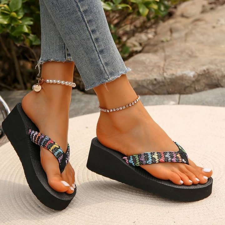 Frau trägt bunte Plateau-Flip-Flops und Jeans, Schmuck an den Knöcheln, auf Steinboden. Sommermode, bequeme Sandalen, trendige Fußbekleidung.