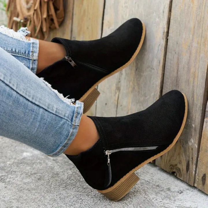Schwarze Damen-Stiefeletten mit Reißverschluss, kombiniert mit zerrissenen Jeans, auf Holzboden. Modische Herbstschuhe, bequeme Absatzstiefeletten.