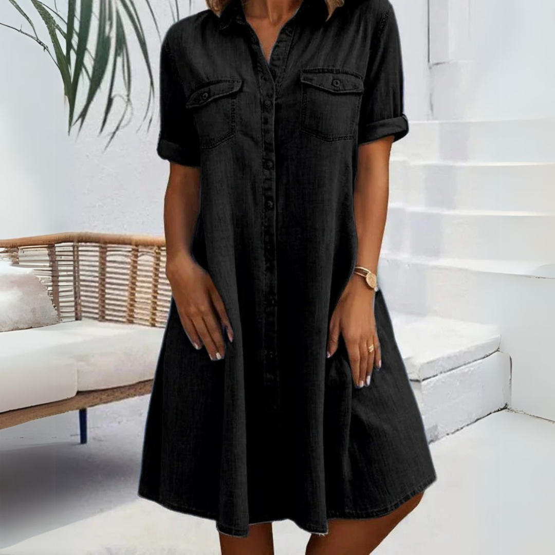 Frau in schwarzem Hemdkleid, knielang, mit kurzen Ärmeln, steht vor weißer Treppe und Rattan-Sofa. Sommermode, Freizeitkleidung, eleganter Look.