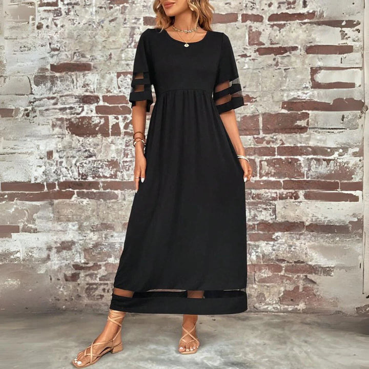 Frau in elegantem, schwarzem Maxikleid mit transparenten Ärmeln vor rustikaler Backsteinwand, trägt Sandalen und Schmuck. Modetrend, Abendmode.