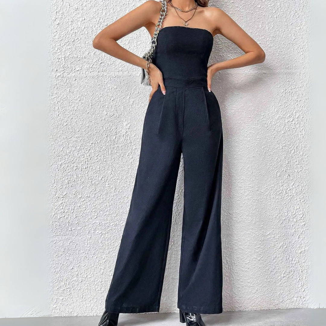 Frau in schwarzem, schulterfreiem Jumpsuit vor weißer Wand, modisch elegant, mit Kettenriemen-Tasche, trendige Damenmode, stilvoller Look.