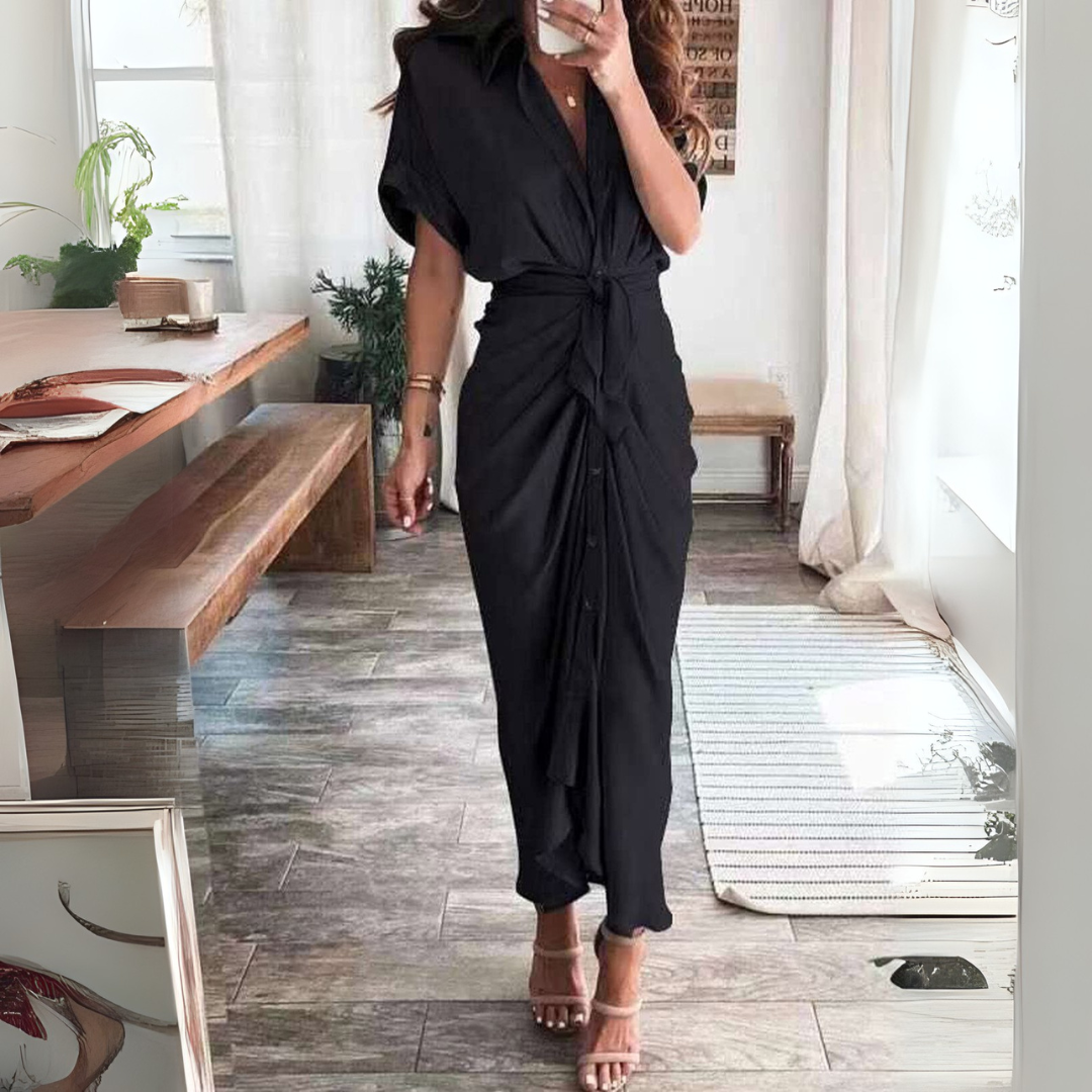 Frau in elegantem schwarzen Maxikleid, steht in modernem Wohnzimmer mit Holztisch und Pflanzen, trägt Sandalen. Stilvolle Mode, Inneneinrichtung.