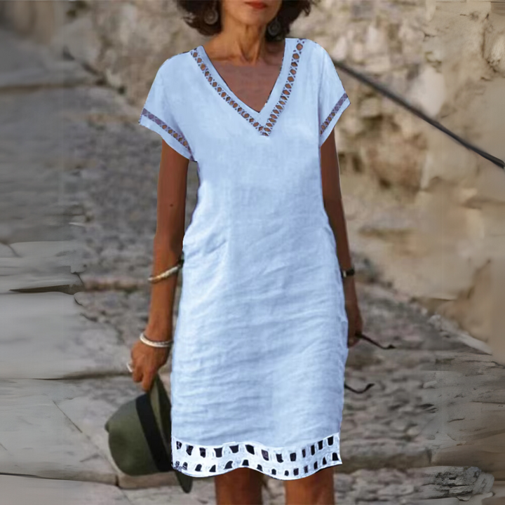 Frau in weißem Sommerkleid mit V-Ausschnitt und Spitzenmuster, hält Hut in der Hand, steht auf gepflastertem Weg. Sommermode, Damenkleidung.