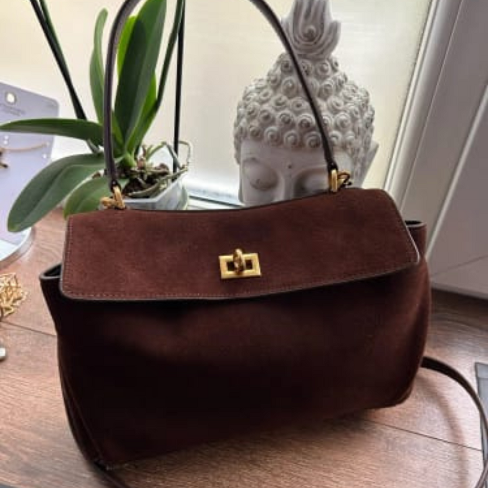 Dunkelbraune Wildlederhandtasche mit goldener Schnalle auf Holztisch, neben grüner Pflanze und Buddha-Statue, elegantes Accessoire.