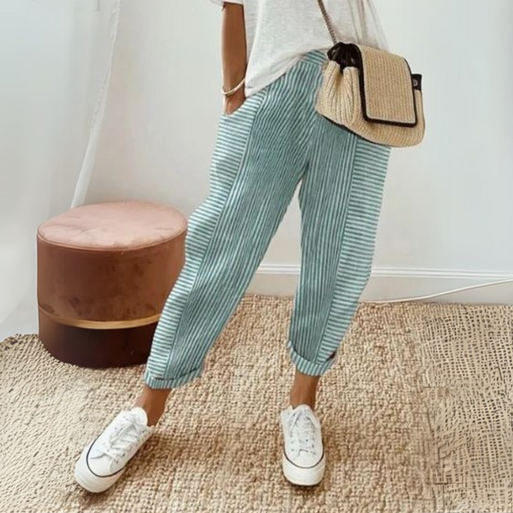 Dame in gestreifter, blauer Hose und weißem T-Shirt, trägt weiße Sneakers und eine Strohtasche. Modetrend, lässiger Sommerlook, Damenmode.