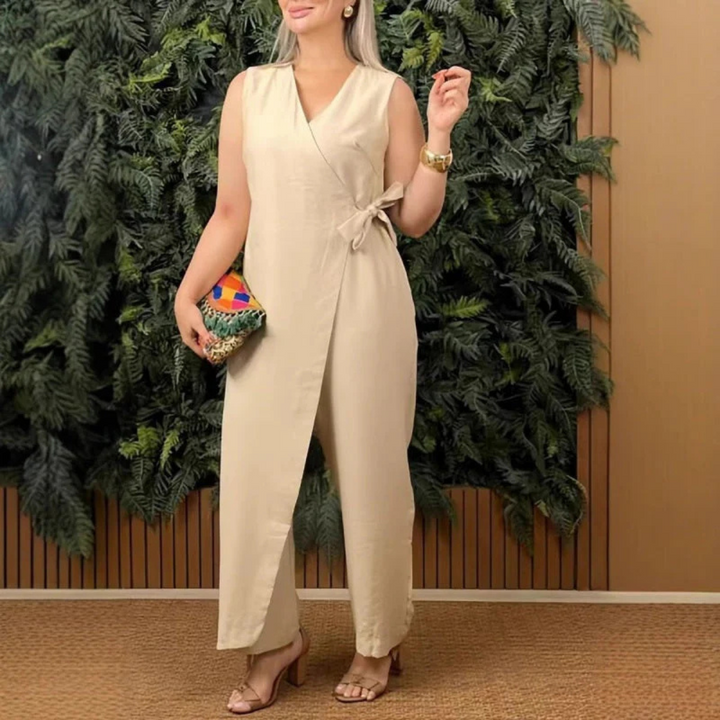 Frau in beigem Jumpsuit mit Wickeldesign, hält bunte Clutch, steht vor grüner Pflanzenwand. Elegante Mode, Sommeroutfit, stilvolle Accessoires.
