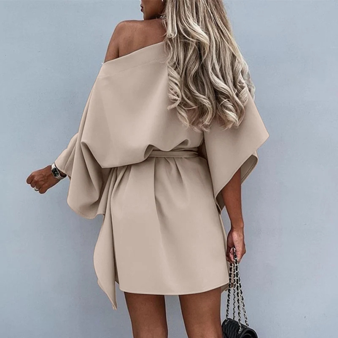 Blonde Frau in beigem, schulterfreiem Kleid mit Gürtel, hält schwarze Kettentasche. Elegantes Modeoutfit, modernes Design, trendiger Look.