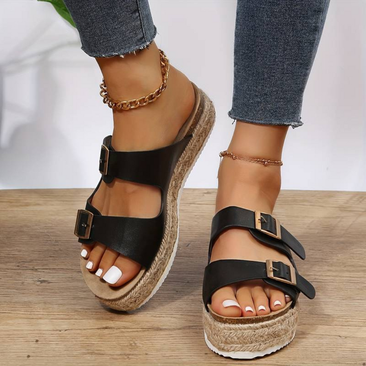 Damenfüße in schwarzen Sandalen mit Plateau und Schnallen, kombiniert mit Jeans und Fußkettchen, auf Holzboden. Modische Sommerschuhe.