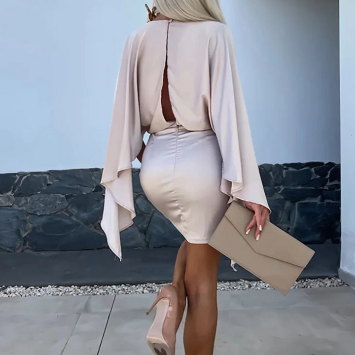 Elegante Frau in beigem Kleid mit Cape-Ärmeln und High Heels, hält eine passende Clutch. Stilvolle Mode, Abendgarderobe, schickes Outfit.