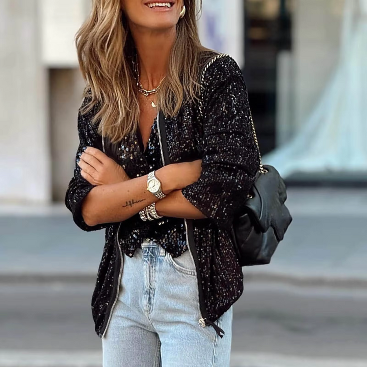 Frau in modischer schwarzer Paillettenjacke, hellblauer Jeans und schwarzer Tasche. Lächelnd, mit Schmuck und Armbanduhr. Straßenhintergrund.