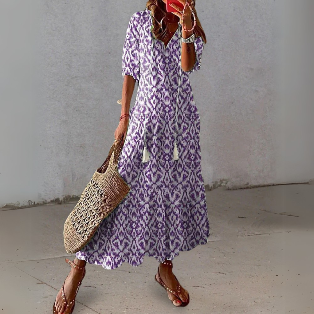 Frau in lila-weißem Boho-Kleid mit Ethno-Muster, hält eine geflochtene Tasche, trägt Sandalen. Sommermode, lässiger Stil, Outdoor.