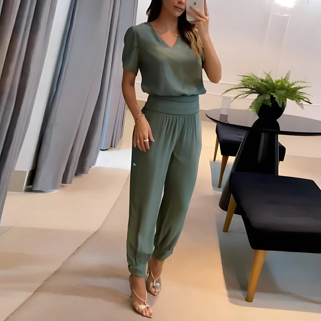 Frau in grünem Jumpsuit macht Selfie im modernen Wohnzimmer, elegante Einrichtung, Pflanzen, Vorhänge, stilvolle Mode, Innenarchitektur.