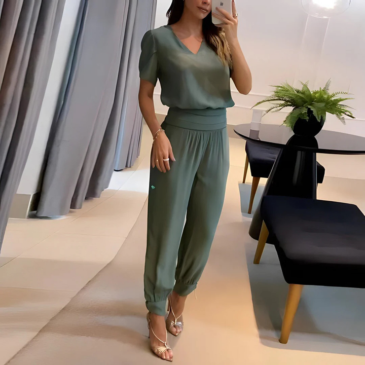 Frau in grünem Jumpsuit macht Selfie im modernen Wohnzimmer, elegante Einrichtung, Pflanzen, Vorhänge, stilvolle Mode, Innenarchitektur.