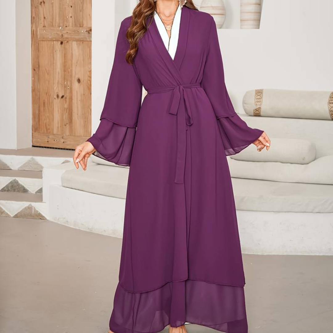 Frau in lila Maxikleid mit Gürtel, stehend in modernem Wohnzimmer. Elegantes Abendkleid, modische Damenbekleidung, stilvolle Inneneinrichtung.
