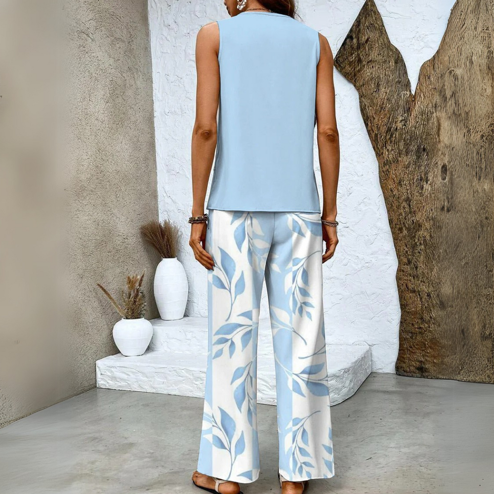 Frau in blauer ärmelloser Bluse und weiß-blauer gemusterter Hose, steht vor rustikaler Wand mit Dekovasen. Elegante Sommermode, lässiger Stil.