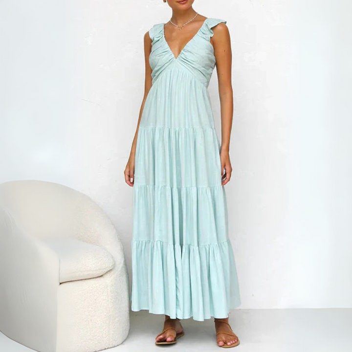 Frau in hellblauem Maxikleid mit Rüschenärmeln steht neben einem weißen Sessel. Sommermode, elegantes Design, Freizeitkleidung.