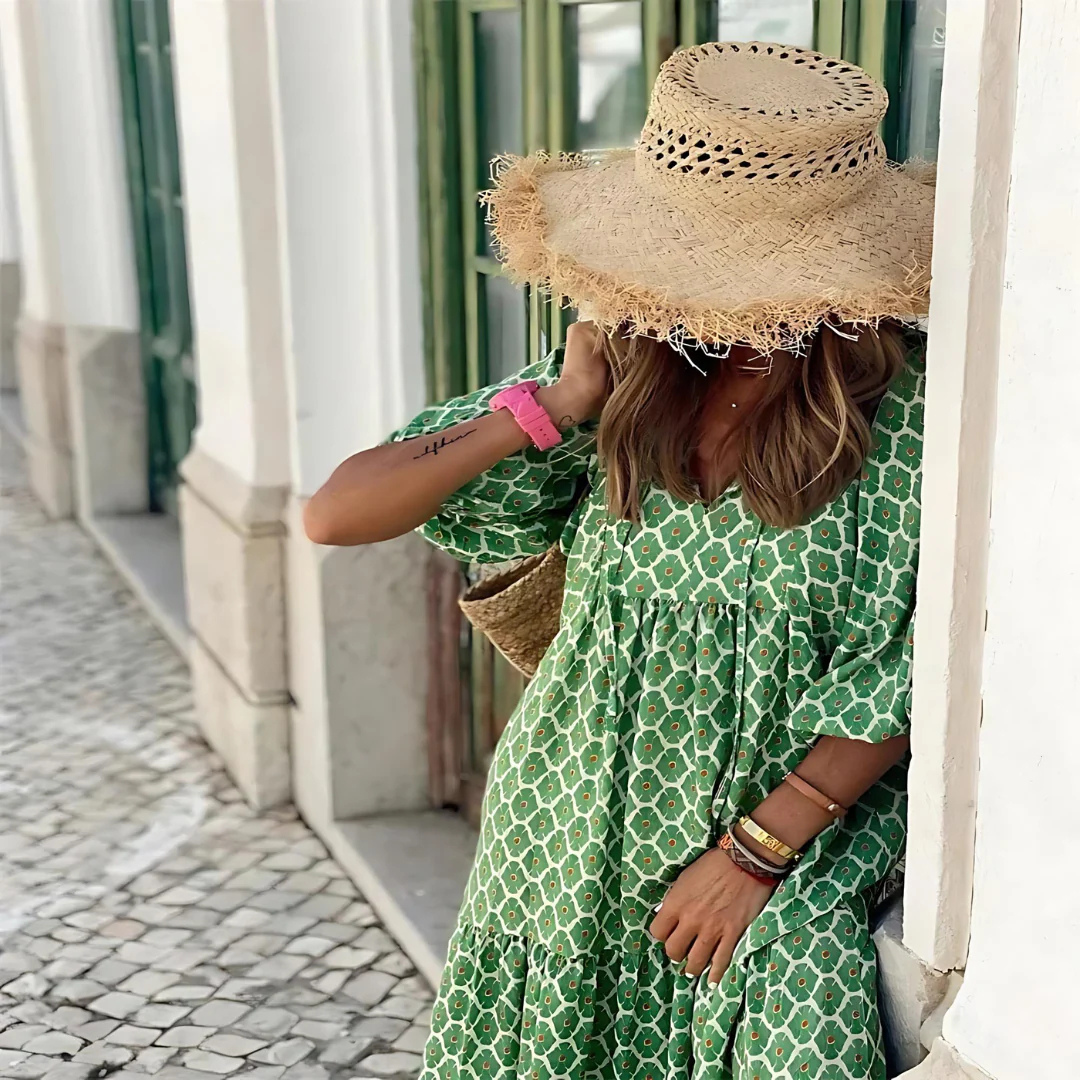 Frau in grünem Kleid und Strohhut lehnt an Wand, Sommermode, Boho-Stil, Stadtbummel, modisches Outfit, entspannter Look, sonniger Tag.