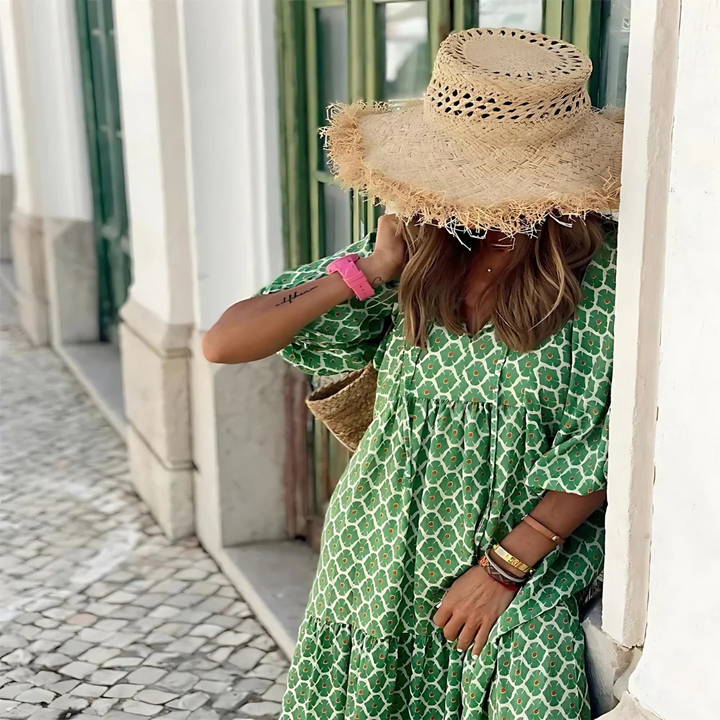 Frau in grünem Kleid und Strohhut lehnt an Wand, Sommermode, Boho-Stil, Stadtbummel, modisches Outfit, entspannter Look, sonniger Tag.