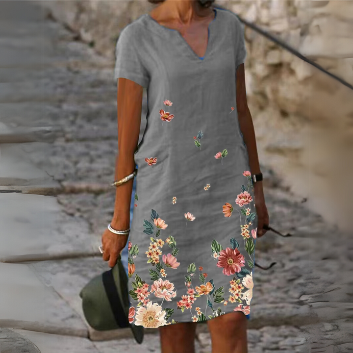 Frau in grauem Kleid mit buntem Blumenmuster, steht auf gepflastertem Weg. Sommermode, lässiger Stil, florales Design, Freizeitkleidung.