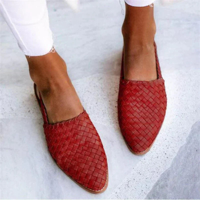 Rote geflochtene Lederslipper auf grauem Boden, getragen von Person in weißen Hosen. Modische Sommerschuhe, handgefertigt, elegant und bequem.