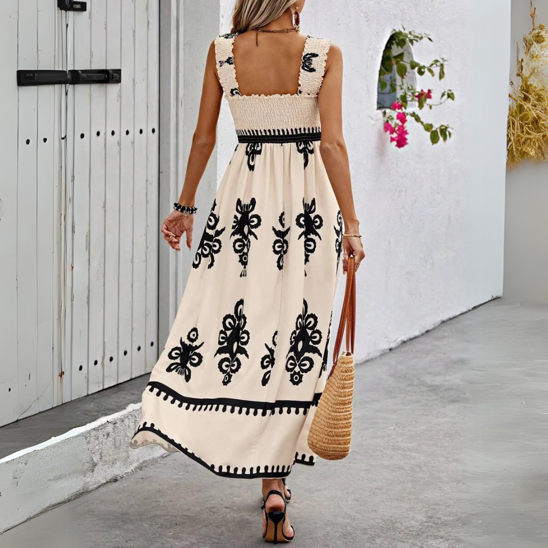 Frau in langem, ärmellosem Sommerkleid mit schwarzem Blumenmuster, trägt Strohtasche, geht auf grauem Gehweg neben weißer Wand mit Blumen.