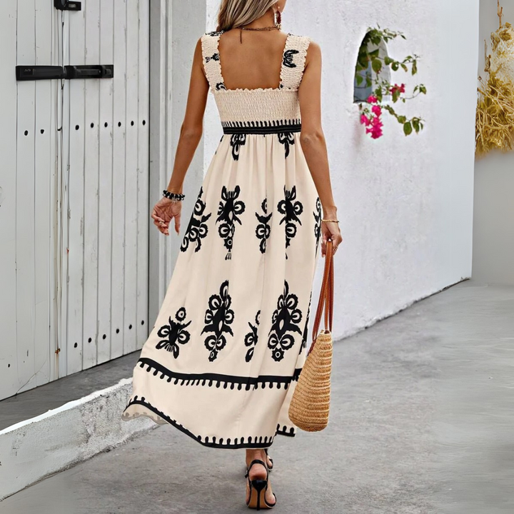 Frau in langem, ärmellosem Sommerkleid mit schwarzem Blumenmuster, trägt Strohtasche, geht auf grauem Gehweg neben weißer Wand mit Blumen.