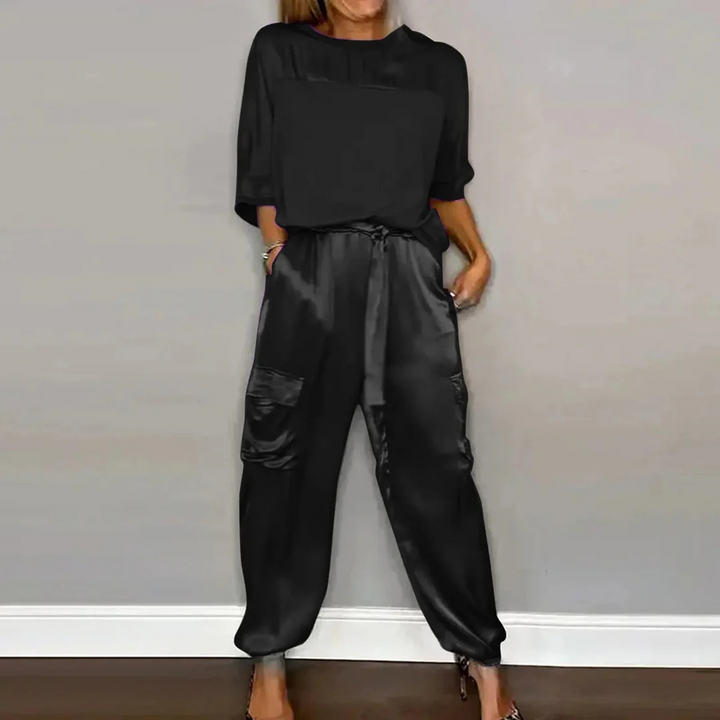 Frau in schwarzem Satin-Overall mit langen Ärmeln und Taschen, steht vor grauer Wand. Modetrend, eleganter Jumpsuit, stilvolle Damenbekleidung.