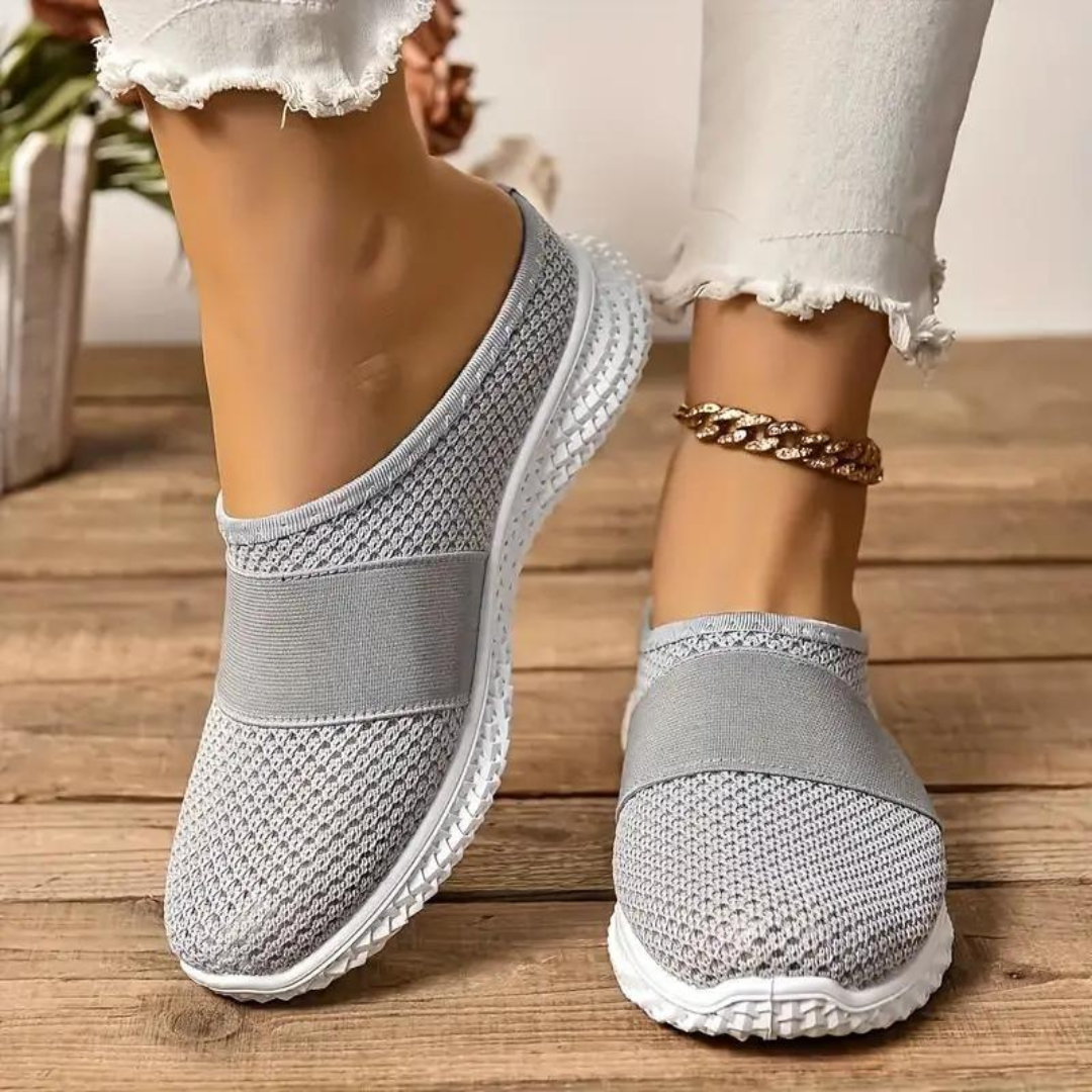 Graue Damen-Sneaker mit Strickmuster und weißer Sohle auf Holzboden, getragen mit weißer Jeans und goldener Fußkette. Bequeme Freizeitschuhe.