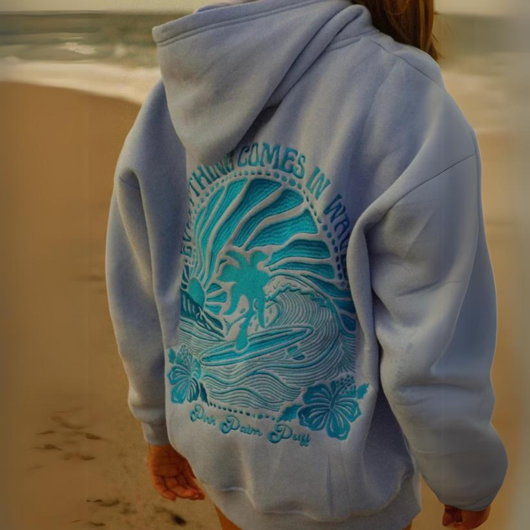 Person am Strand in einem blauen Hoodie mit tropischem Design und Surfer-Motiv. Perfekt für Strandmode, Surfkleidung, Freizeitbekleidung.