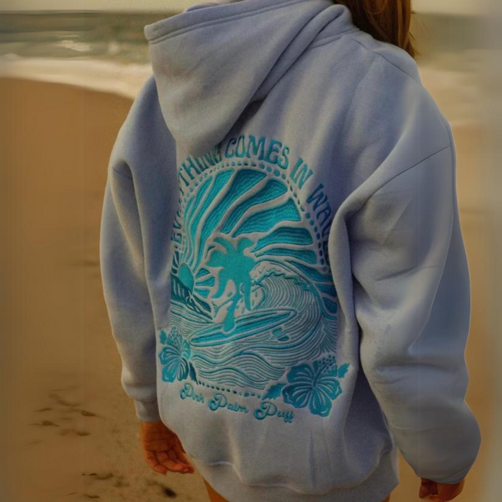 Person am Strand in einem blauen Hoodie mit tropischem Design und Surfer-Motiv. Perfekt für Strandmode, Surfkleidung, Freizeitbekleidung.