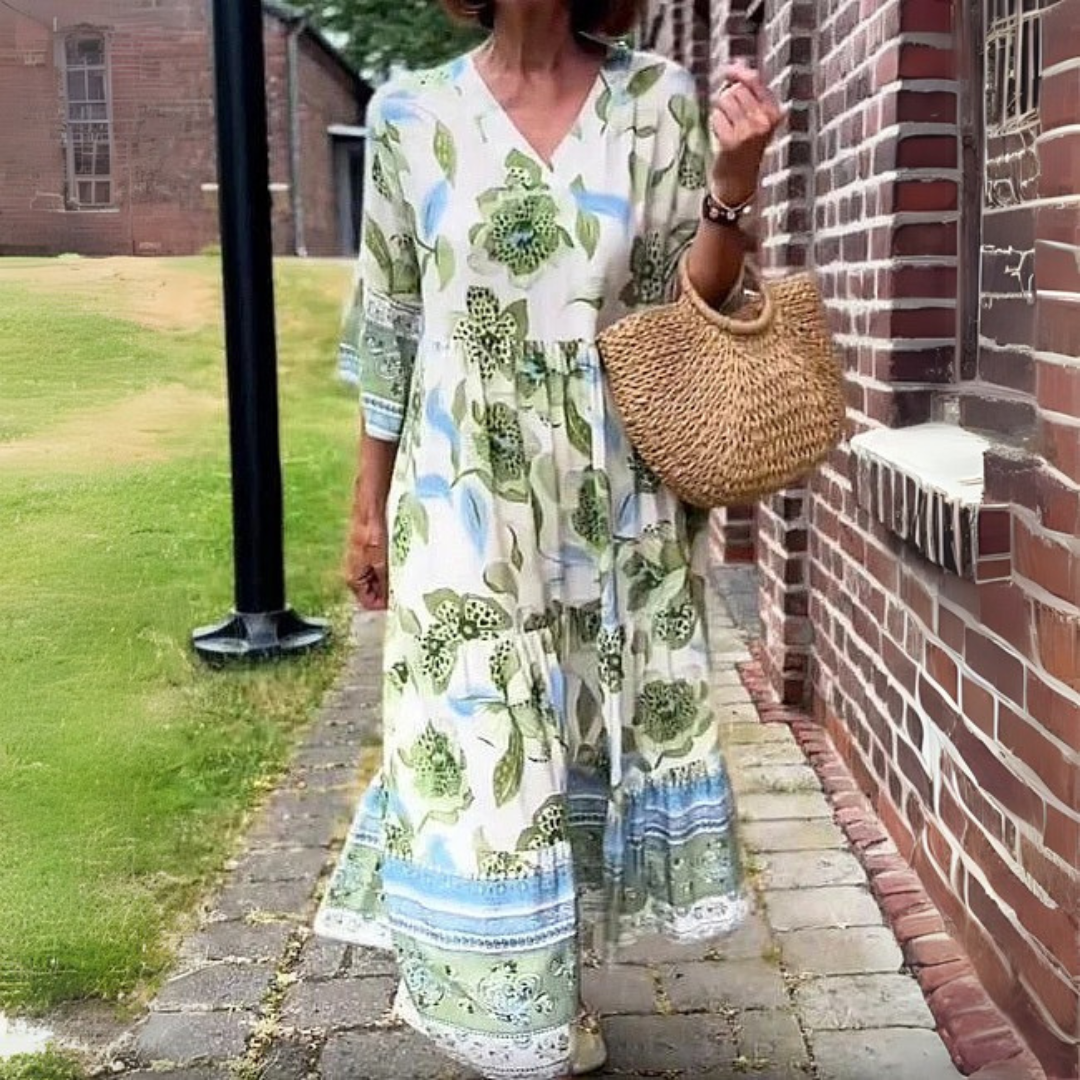 Frau in langem, geblümtem Sommerkleid mit Strohtasche spaziert auf gepflastertem Weg neben Backsteinmauer. Elegante Mode, Sommerkleid, Strohtasche.
