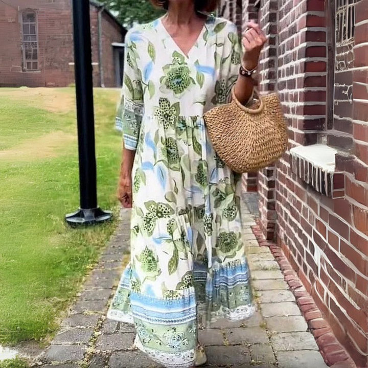 Frau in langem, geblümtem Sommerkleid mit Strohtasche spaziert auf gepflastertem Weg neben Backsteinmauer. Elegante Mode, Sommerkleid, Strohtasche.