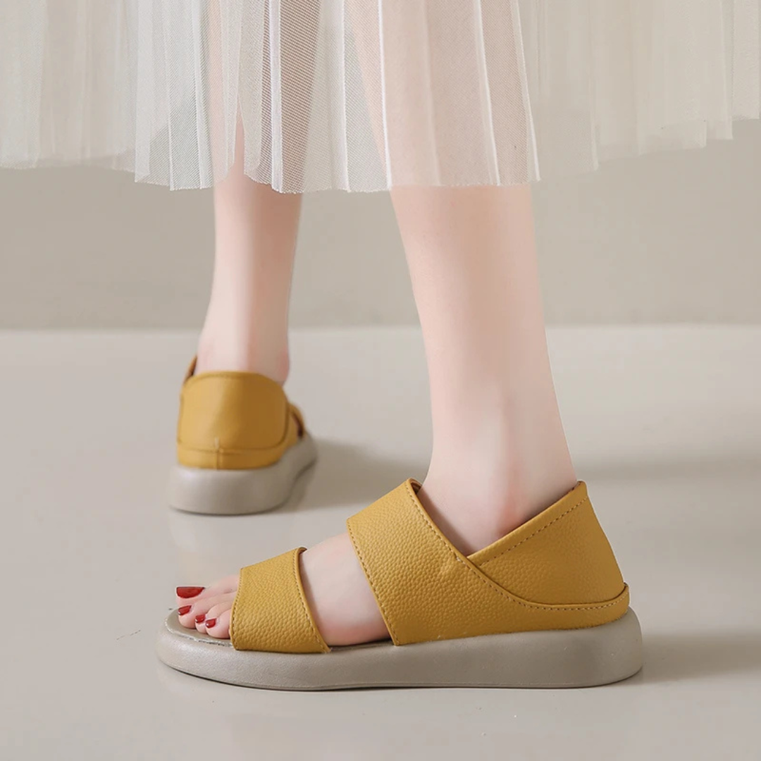 Gelbe Sandalen mit breiten Riemen an Frauenfüßen auf hellem Boden, kombiniert mit einem weißen, plissierten Rock. Ideal für Sommermode und Komfort.