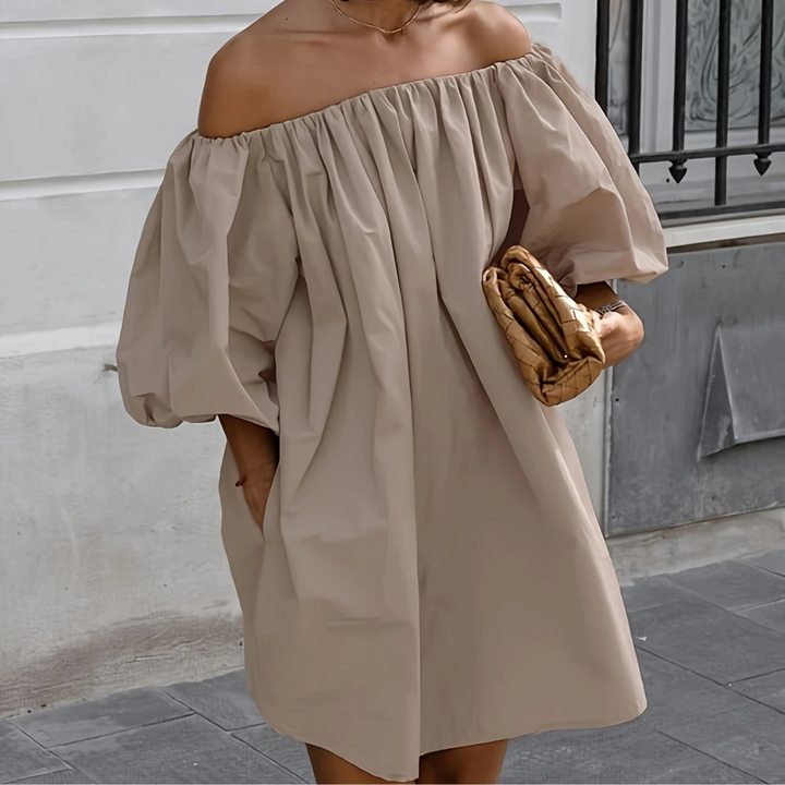 Frau in schulterfreiem, beigem Kleid mit Puffärmeln hält eine gefaltete Tasche. Elegante Sommermode, urbaner Hintergrund.