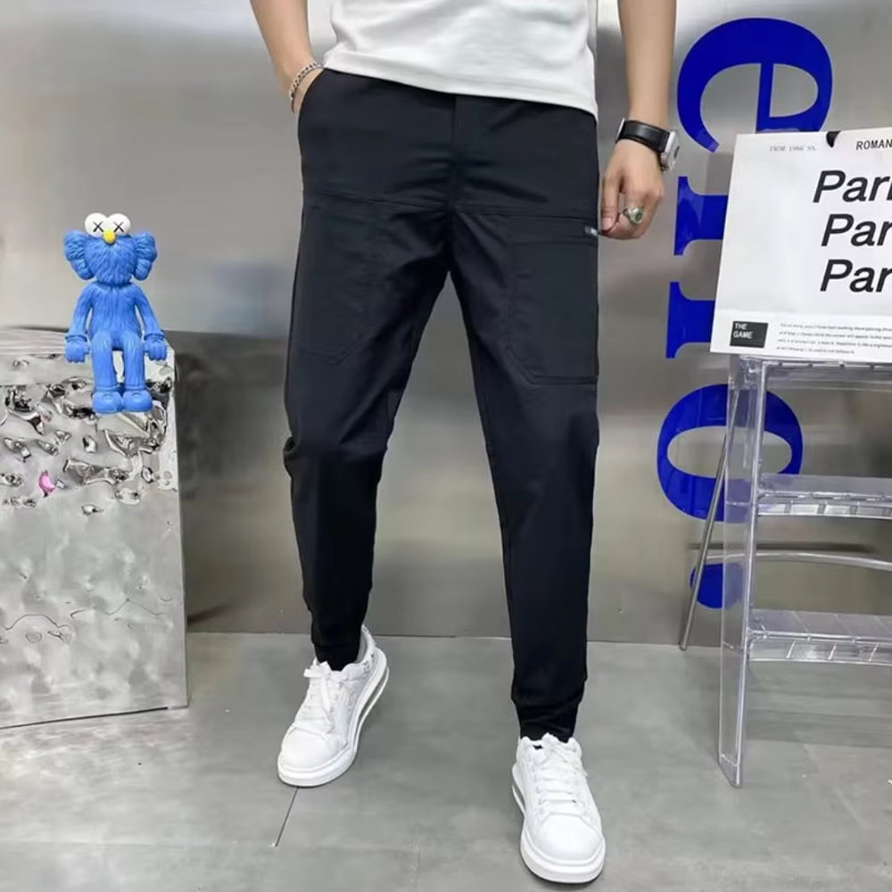 Mann in schwarzer Jogginghose und weißen Sneakers, steht neben blauem Plüschtier und transparentem Schild. Moderne Herrenmode, lässiger Stil.