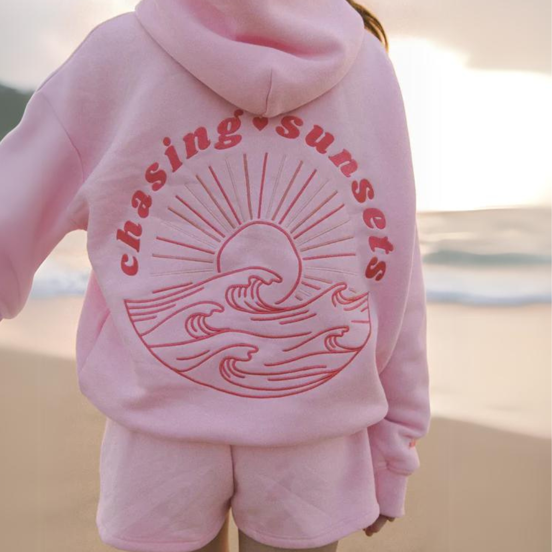 Person in rosa Hoodie mit "chasing sunsets" und Sonnenuntergangs-Design am Strand. Bequeme Strandmode, lässiger Stil, Sonnenuntergang genießen.