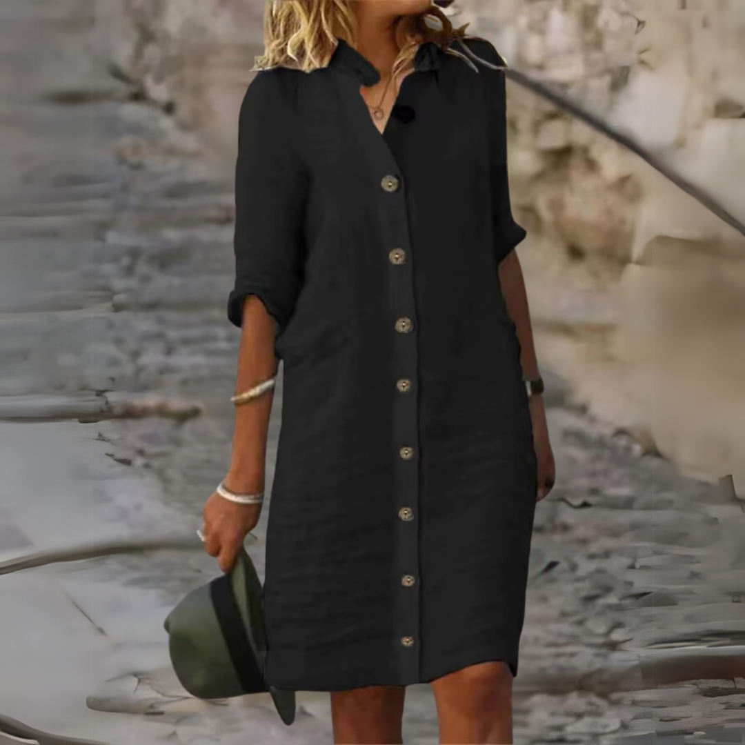 Frau in schwarzem Hemdkleid mit Knopfleiste, hält grünen Hut, steht auf gepflastertem Weg. Modetrend, lässiger Stil, Sommeroutfit.