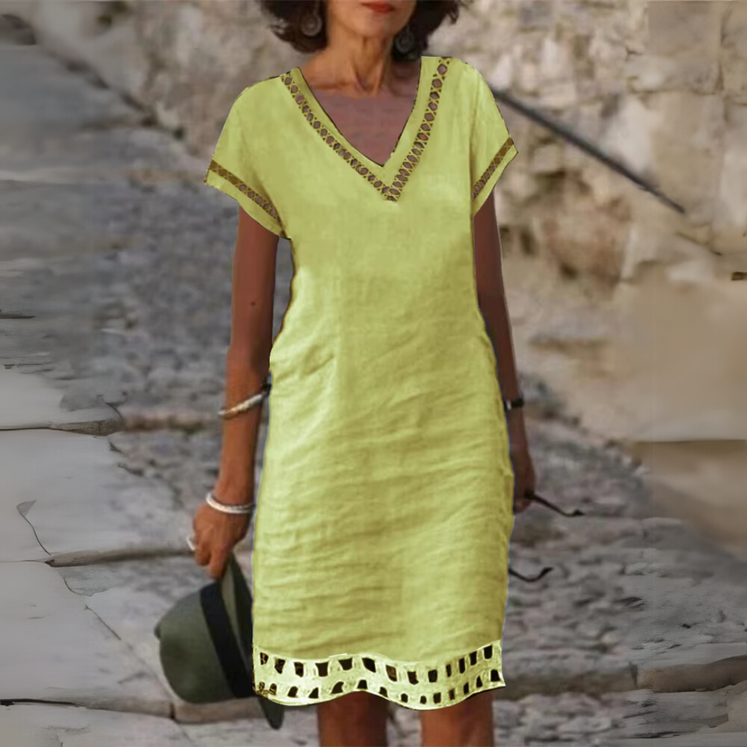 Frau in gelbem Sommerkleid mit V-Ausschnitt und Spitzenverzierung, hält Hut in der Hand, steht auf gepflastertem Weg vor Steinmauer.