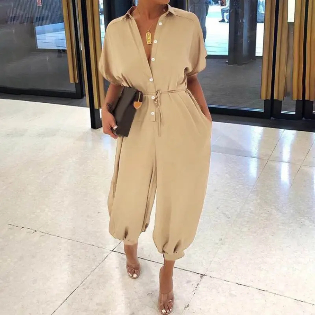 Beige Jumpsuit mit Knopfleiste und Gürtel, kombiniert mit schwarzen High Heels und Clutch, getragen von einer Frau in einem modernen Innenraum.