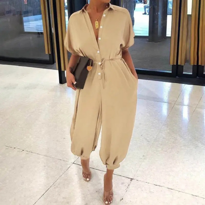 Beige Jumpsuit mit Knopfleiste und Gürtel, kombiniert mit schwarzen High Heels und Clutch, getragen von einer Frau in einem modernen Innenraum.