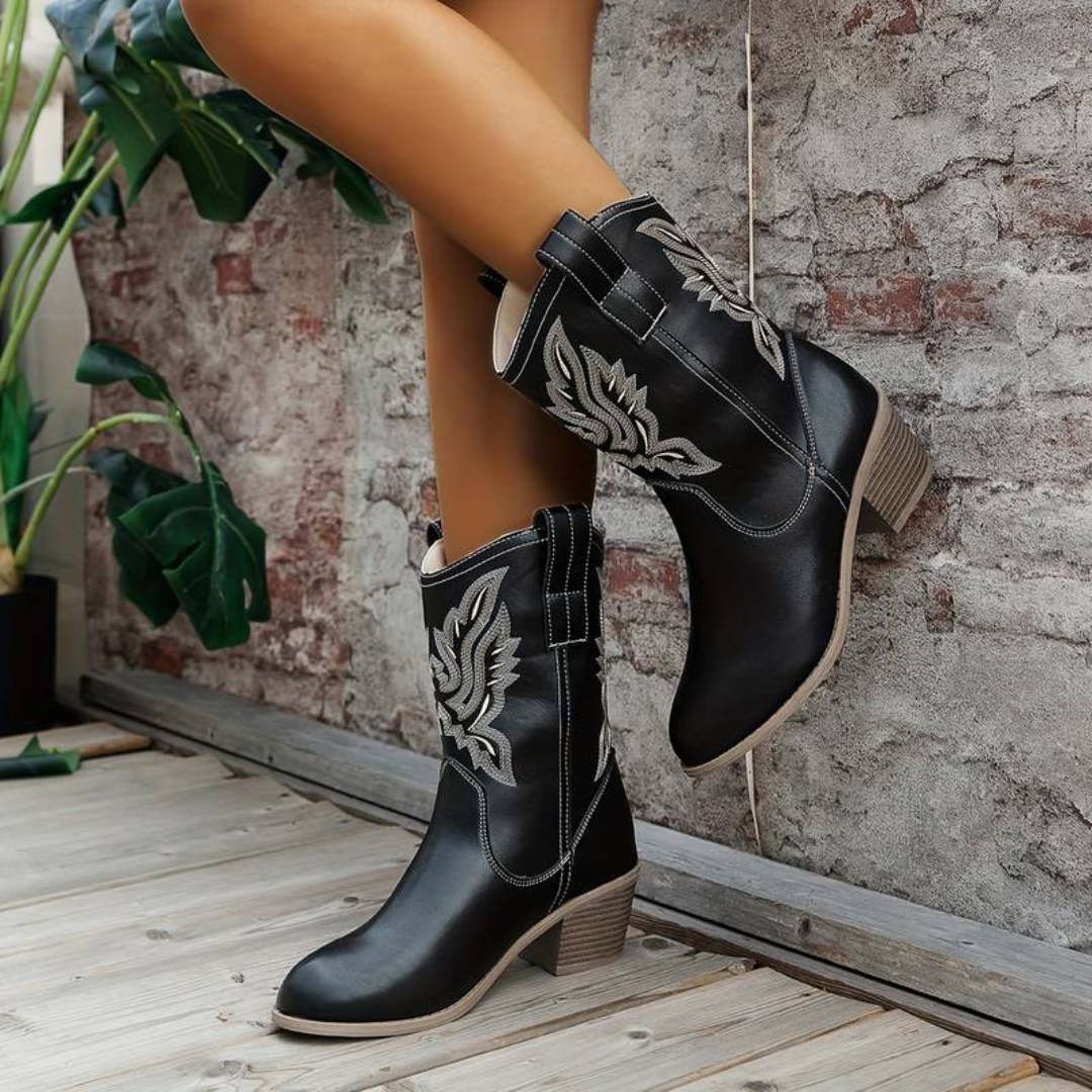 Schwarze Cowboystiefel mit weißen Stickereien, Absatz und runder Spitze, vor einer Ziegelwand. Modische Damenstiefel, Westernstil.