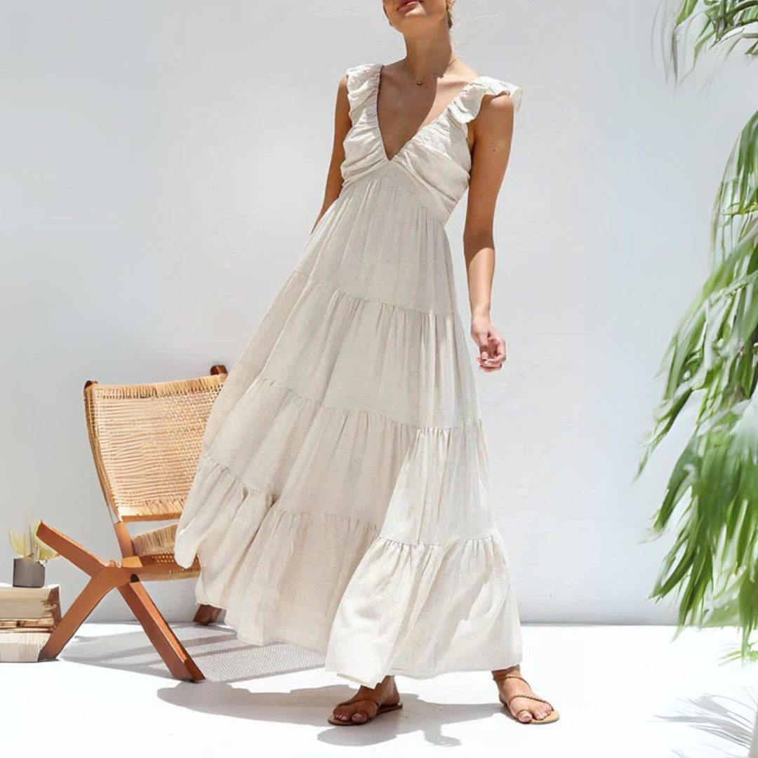 Frau in weißem Sommerkleid mit Rüschen, steht neben einem Rattan-Stuhl. Luftiges Maxikleid, ideal für Strandmode und Sommeroutfits.